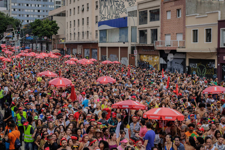 A imagem mostra uma grande multidão reunida em uma rua， com várias pessoas segurando guarda-sóis vermelhos. O ambiente é urbano， com prédios ao fundo e grafites nas paredes. A maioria das pessoas está vestida de forma colorida， sugerindo um evento festivo. Algumas pessoas usam fantasias e acessórios， e há também pessoas com coletes de segurança visíveis.