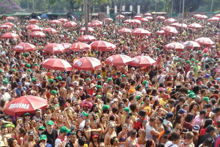 A imagem mostra uma grande multidão reunida em um evento ao ar livre， com muitas pessoas usando roupas coloridas e acessórios festivos. Há várias sombrinhas vermelhas abertas， criando um padrão visual. A cena é animada， com pessoas dançando e se divertindo sob a luz do sol， em um ambiente festivo.