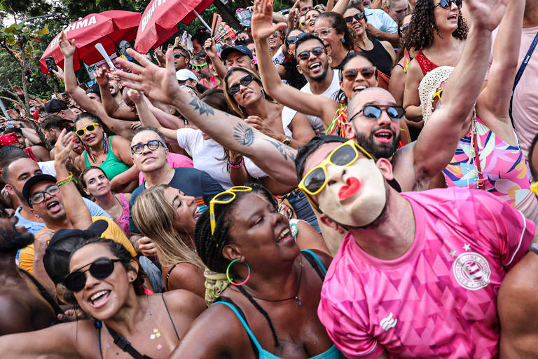 A imagem mostra uma multidão animada durante uma festa， possivelmente um carnaval. As pessoas estão sorrindo， dançando e se divertindo. Algumas usam óculos de sol e roupas coloridas， enquanto uma pessoa se destaca usando uma máscara com lábios vermelhos. Ao fundo， há guarda-sóis vermelhos e uma atmosfera festiva.