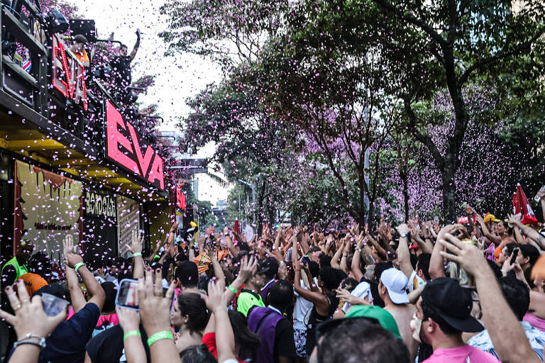 A imagem mostra uma multidão animada em uma festa ao ar livre， com pessoas levantando as mãos. Confetes coloridos estão caindo do céu， criando um ambiente festivo. Ao fundo， há um edifício com a palavra 039;EVA039; em destaque. A cena é cercada por árvores e um clima de celebração.