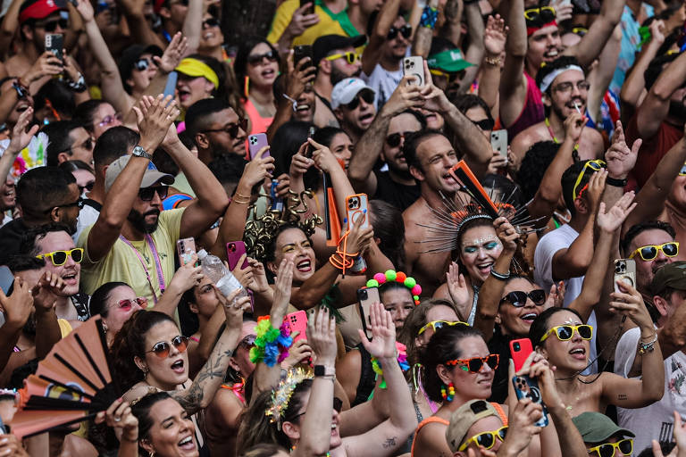 A imagem mostra uma grande multidão em um evento festivo, com pessoas sorrindo e levantando as mãos. Muitas delas usam óculos de sol coloridos e seguram celulares, algumas com acessórios coloridos na cabeça. O ambiente é vibrante e alegre, refletindo um clima de celebração.