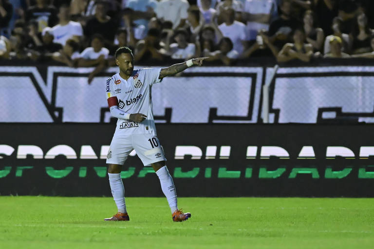 Um jogador de futebol， vestindo um uniforme branco com detalhes em preto e a camisa número 10， está em campo apontando para a frente. Ele usa uma faixa de capitão no braço. Ao fundo， há uma multidão de torcedores， e uma faixa preta com a palavra 039;RESPONSABILIDADE039; em letras grandes e brancas.