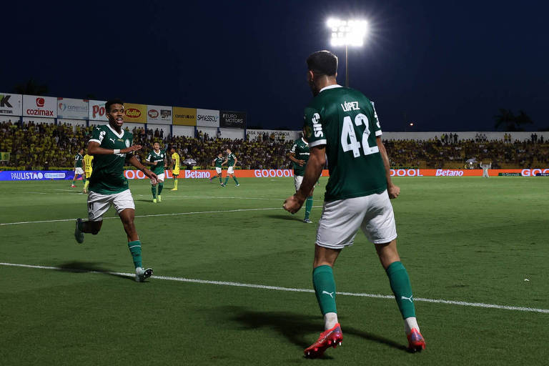 Dois jogadores de futebol estão se celebrando em um campo iluminado por refletores à noite. Um jogador está correndo em direção ao outro， que está levantando os braços em comemoração. Ambos estão vestindo uniformes verdes， com um deles usando a camisa número 42. Ao fundo， há uma multidão assistindo ao jogo.