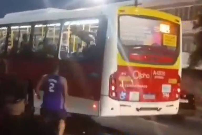 A imagem mostra um ônibus urbano em movimento， com a parte traseira visível. O ônibus é de cor branca com detalhes em vermelho e amarelo. Há pessoas visíveis dentro do ônibus. Na parte inferior da imagem， um homem vestido com uma camiseta roxa e o número 2 nas costas está correndo em direção ao ônibus. 