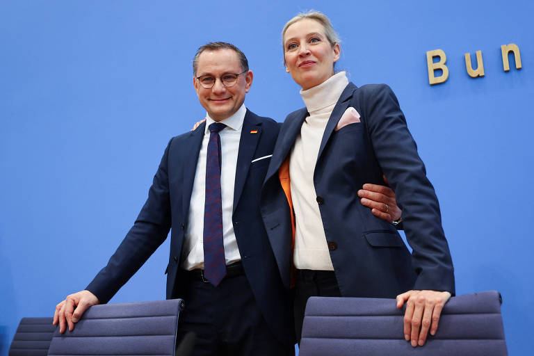 Duas pessoas estão posando para a foto em um ambiente formal. O homem à esquerda está usando um terno escuro com uma gravata e sorrindo. A mulher à direita está vestindo um blazer escuro e uma blusa clara， também sorrindo. Ao fundo， há uma parede azul com a palavra 039;Bundes039; visível.