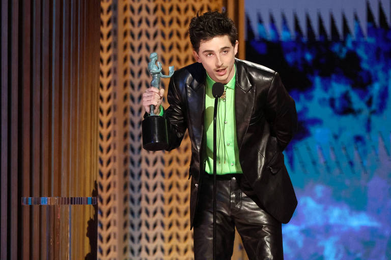 Um jovem homem está em um palco， segurando um troféu em uma mão e um microfone na outra. Ele usa uma camisa verde-limão e um terno preto brilhante. O fundo apresenta um padrão geométrico em tons de marrom e azul.