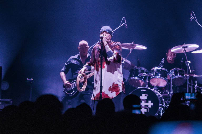 A imagem mostra um cantor em um palco durante um show ao vivo， segurando um instrumento de percussão. Ele está vestido com uma camisa de cores claras e escuras， com manchas vermelhas. Ao fundo， há um músico tocando baixo e outro na bateria. A iluminação é suave， criando um ambiente de performance musical.