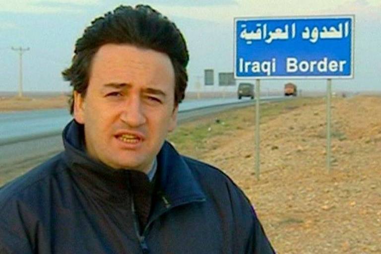 Um repórter está em pé em uma estrada， com um sinal ao fundo que diz 039;Iraqi Border039; em inglês e árabe. O cenário é desértico， com uma estrada visível e algumas estruturas ao longe. O repórter está usando um casaco escuro e parece estar falando para a câmera.