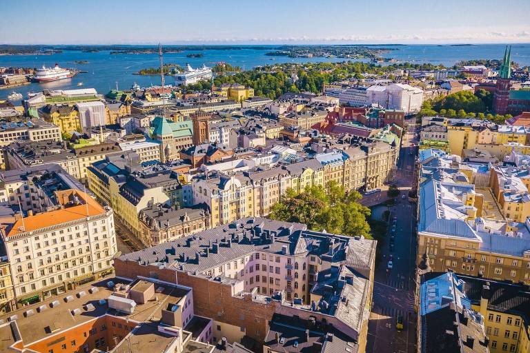 Imagem aérea da cidade de Helsinque， na Finlândia， mostrando uma vista panorâmica do centro urbano. É possível ver edifícios de diferentes cores e estilos arquitetônicos， ruas e áreas verdes. Ao fundo， há o mar e algumas ilhas， além de navios ancorados.