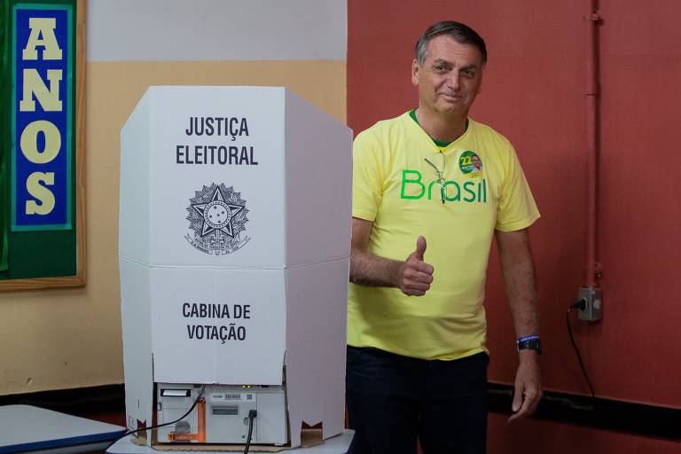 Um homem está ao lado de uma cabine de votação, fazendo um sinal de positivo. Ele usa uma camiseta amarela com a palavra 'Brasil' em verde. A cabine de votação é branca e possui as inscrições 'JUSTIÇA ELEITORAL' e 'CABINA DE VOTAÇÃO'. Ao fundo, há uma parede com um quadro verde e a palavra 'ANOS' em letras grandes e coloridas.