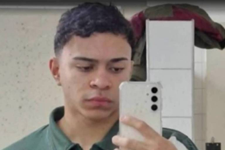 Um jovem está tirando uma selfie em um espelho. Ele usa uma camiseta verde com um emblema no lado esquerdo do peito. O fundo mostra uma parede clara e um objeto verde pendurado.