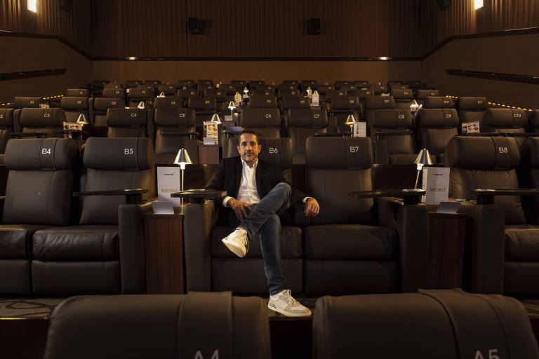 Um homem está sentado em uma poltrona de cinema em um auditório vazio. Ele usa uma jaqueta preta e calças jeans, com tênis brancos. O ambiente é composto por várias poltronas reclináveis, todas numeradas, com pequenas lâmpadas ao lado. O fundo é escuro, com paredes de madeira e iluminação suave.