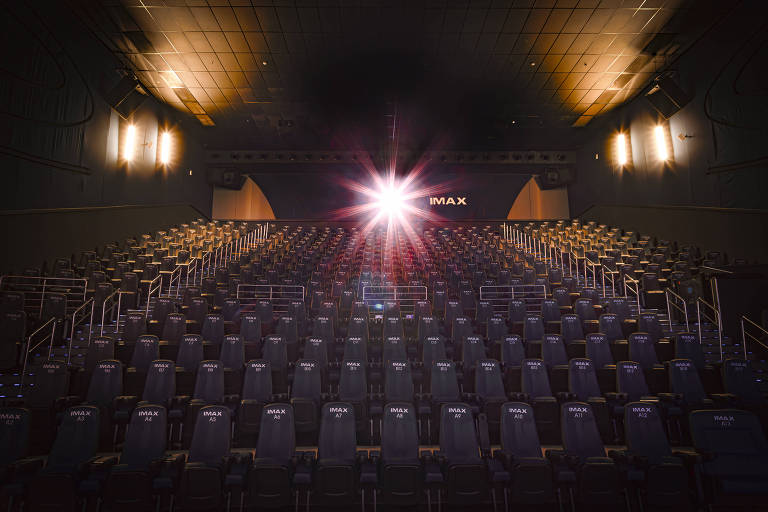 A imagem mostra uma sala de cinema IMAX vazia, com fileiras de assentos escuros organizados em um ângulo. No fundo, há uma tela grande com a palavra 'IMAX' iluminada. Um feixe de luz brilha a partir da tela, criando um efeito visual dramático. O teto é alto e há luzes no ambiente, destacando a estrutura da sala.