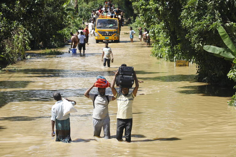 A imagem mostra uma estrada inundada， com pessoas caminhando na água até a cintura. Dois homens estão carregando bagagens， um com uma mochila e outro com roupas. Ao fundo， um ônibus amarelo está parado， e várias outras pessoas estão visíveis na estrada， algumas também carregando objetos. A vegetação ao redor é densa， com árvores e plantas.