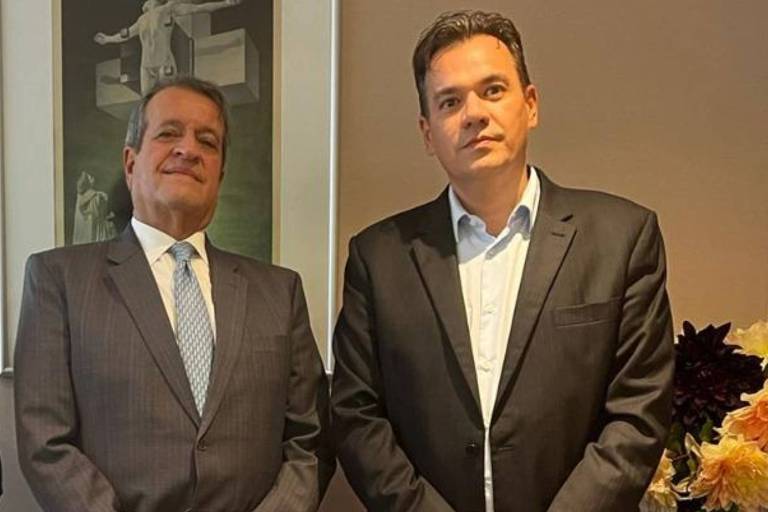 Três homens estão posando para a foto em um ambiente interno. O homem à esquerda está vestindo uma camisa escura e um blazer， o do meio está usando um terno cinza e uma gravata clara， e o da direita está com uma camisa clara e um blazer escuro. Ao fundo， há uma obra de arte na parede e um arranjo de flores em uma mesa.