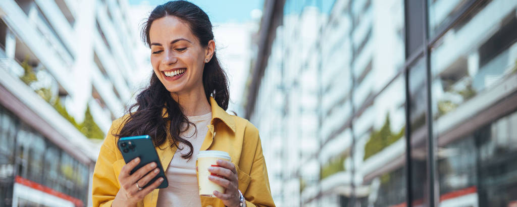 Uma mulher com cabelo longo e ondulado, vestindo uma camisa clara e um casaco amarelo, está sorrindo enquanto olha para o celular que segura com uma mão. Na outra mão, ela segura um copo de café. Ao fundo, há edifícios modernos refletidos em uma superfície de vidro.