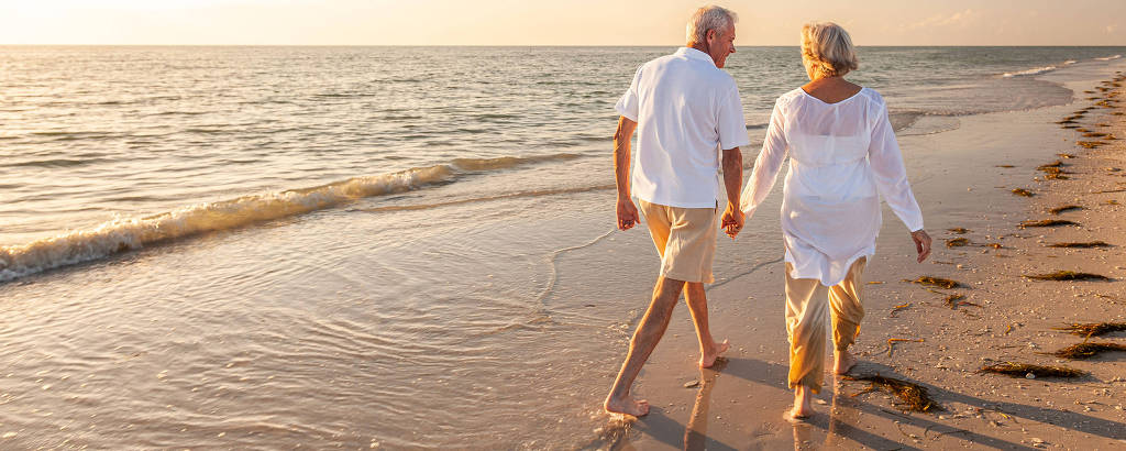 Um casal de idosos caminha de mãos dadas na praia, com o mar ao fundo. Eles estão descalços na areia molhada, e a luz do sol ilumina a cena, criando um ambiente tranquilo e romântico. O homem usa uma camisa branca e calças claras, enquanto a mulher veste uma blusa branca e calças claras também.