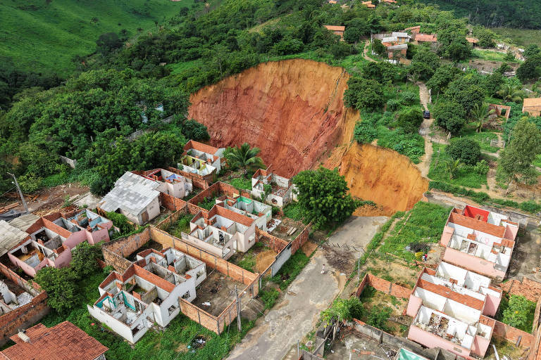 A imagem mostra uma vista aérea de uma área urbana afetada por um deslizamento de terra. À esquerda， há várias casas com telhados de cerâmica e paredes em diferentes estados de conservação. À direita， um grande deslizamento de terra expõe uma encosta de solo avermelhado， cercada por vegetação. O terreno ao redor é predominantemente verde， com árvores e algumas casas visíveis ao fundo.