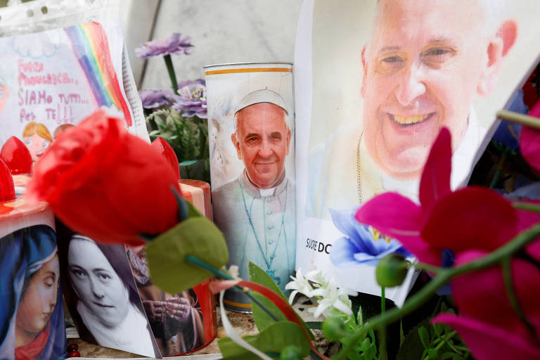 A imagem mostra um altar com velas， flores e várias fotos. Entre as fotos， há uma imagem do Papa Francisco e outras figuras religiosas. Um vaso com uma rosa vermelha está em destaque， e há também mensagens escritas em papel ao fundo.