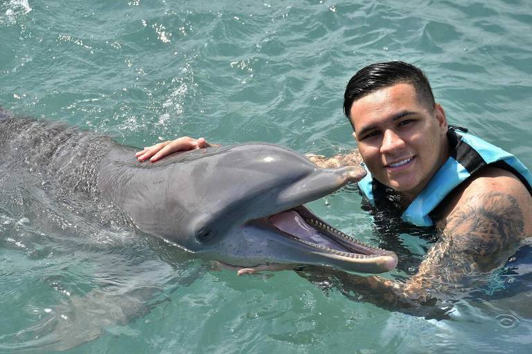 Um homem sorridente está na água, posando ao lado de um golfinho. O golfinho está com a boca aberta, parecendo feliz, enquanto o homem acaricia sua cabeça. Ambos estão em águas claras e tranquilas, com luz do sol refletindo na superfície.