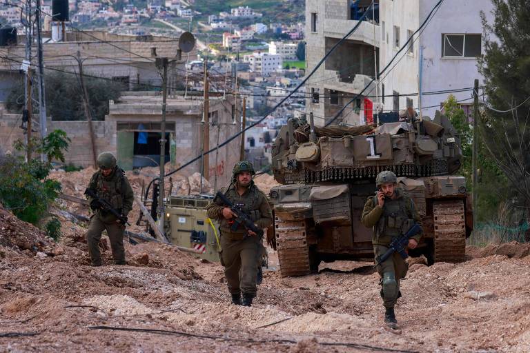 A imagem mostra soldados caminhando em uma área urbana， com um tanque de guerra ao fundo. Os soldados estão vestidos com uniformes militares e portando armas. O cenário é montanhoso， com edifícios e casas visíveis ao fundo， além de fios elétricos e vegetação ao lado da estrada de terra.