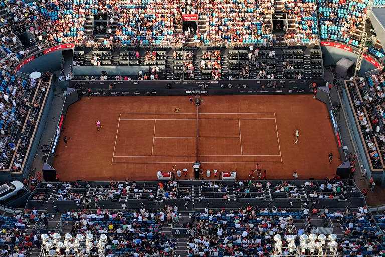 Imagem aérea de um estádio de tênis com uma quadra de saibro no centro. O estádio está cheio de espectadores， com assentos em várias camadas ao redor da quadra. Dois jogadores estão em ação na quadra， um à esquerda e outro à direita. O ambiente é vibrante， com muitas pessoas assistindo ao jogo.