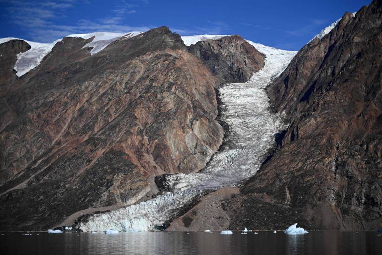 A imagem mostra uma grande montanha com um glaciar descendo por sua encosta. O glaciar é visível na parte inferior da montanha， onde se encontra com a água. O céu está claro e azul， e há algumas áreas de neve no topo da montanha.