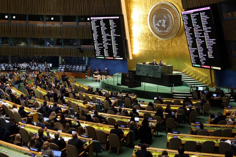 A imagem mostra a Assembleia Geral da Organização das Nações Unidas (ONU) em uma sessão. O ambiente é amplo， com várias fileiras de assentos ocupados por delegados. No centro， há uma mesa com representantes， e ao fundo， uma grande parede dourada com o emblema da ONU. Telas digitais exibem informações， e o espaço é iluminado de forma a destacar a importância do evento.