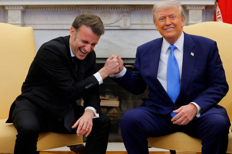 Na imagem， duas pessoas estão sentadas em cadeiras em um ambiente formal. À esquerda， um homem de terno escuro e gravata clara está sorrindo e rindo， enquanto à direita， um homem de terno azul claro e gravata azul escura também está sorrindo. Eles estão se cumprimentando com um aperto de mão， em um gesto de camaradagem. Ao fundo， há uma lareira e uma decoração clássica.