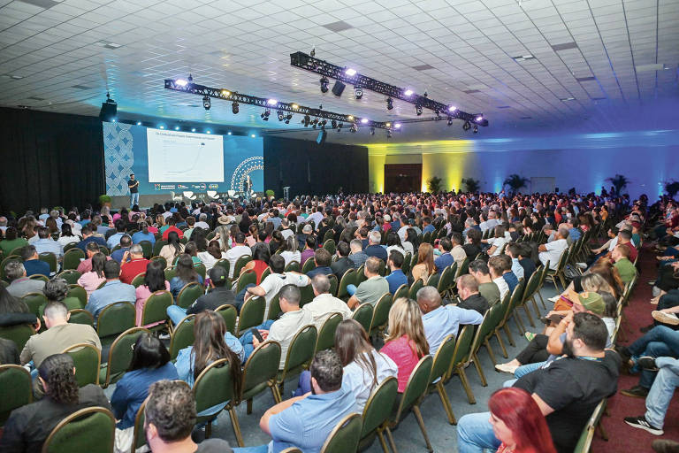 A imagem mostra um grande auditório cheio de pessoas assistindo a uma apresentação. Há uma tela grande à frente， onde um apresentador está falando. O público é composto por homens e mulheres， muitos dos quais estão sentados em cadeiras verdes. O ambiente é bem iluminado， com luzes coloridas ao fundo.