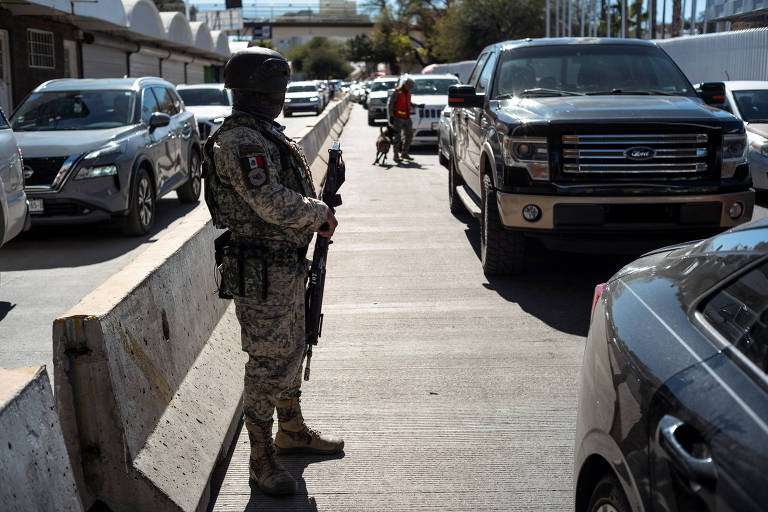 A imagem mostra um soldado armado em um posto de controle， em pé ao lado de uma barreira de concreto. O soldado está usando um uniforme camuflado e um capacete， segurando uma arma. Ao fundo， há uma fila de veículos， incluindo caminhonetes e SUVs， em uma estrada. A cena parece ser em uma área urbana， com edifícios visíveis ao fundo.