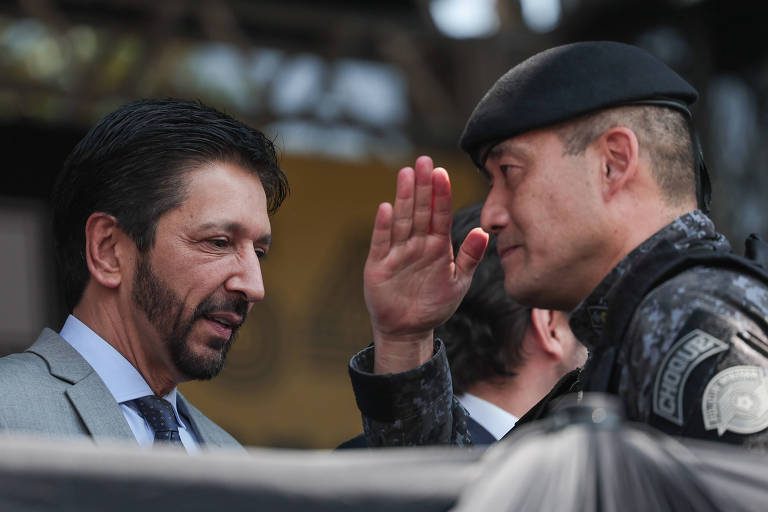 A imagem mostra um homem de terno conversando com um policial. O homem tem cabelo escuro e barba， enquanto o policial está usando um uniforme preto e um boné. O policial está fazendo um gesto de saudação com a mão. Ao fundo， há uma estrutura que parece ser um evento ao ar livre.