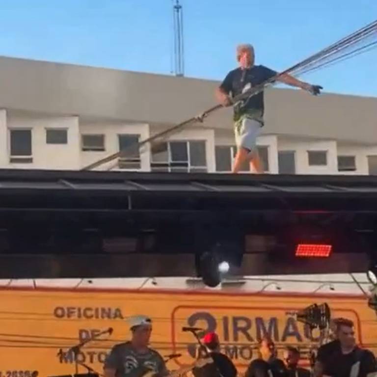 Um artista está em cima de um palco， equilibrando-se enquanto segura um cabo. Ele usa uma camiseta preta e shorts. Ao fundo， há uma banda tocando， com músicos visíveis. O cenário é urbano， com um prédio e fios elétricos ao fundo. A iluminação do palco é visível， com luzes vermelhas acesas.