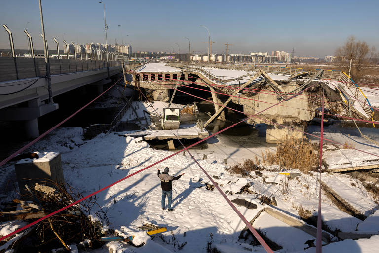 A imagem mostra uma ponte danificada， com partes dela colapsadas e caídas em um terreno coberto de neve. Um homem está de pé， com os braços levantados， em frente à estrutura destruída. Fios e cabos estão espalhados pela área， e há uma barreira de segurança ao fundo. O céu está claro e há edifícios ao longe.