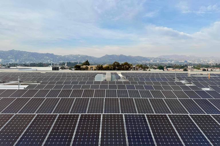 A imagem mostra uma grande área coberta por painéis solares em um telhado， com uma vista panorâmica de uma cidade ao fundo. O céu está parcialmente nublado， e as montanhas são visíveis na distância.