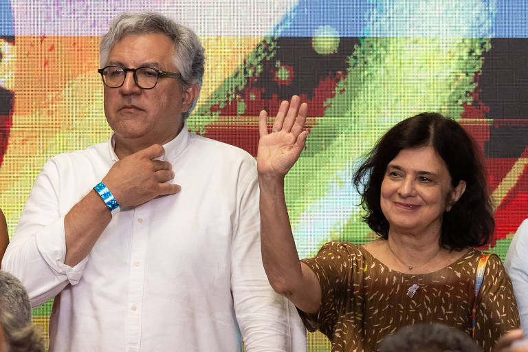 A imagem mostra duas pessoas em um evento. À esquerda， um homem com cabelo grisalho e óculos， vestindo uma camisa branca e uma pulseira azul. Ele está com a mão no peito. À direita， uma mulher com cabelo escuro e liso， usando uma blusa dourada. Ela está acenando com a mão. Ao fundo， há uma tela colorida com padrões vibrantes.