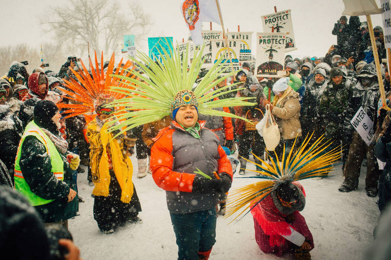 A imagem mostra um grupo de pessoas reunidas em um protesto durante uma nevasca. No centro， duas crianças vestindo roupas coloridas e adornos de penas dançam. Uma delas usa um casaco laranja e a outra um traje com penas verdes e amarelas. Ao fundo， há uma multidão de manifestantes segurando cartazes que dizem 039;PROTEJA A ÁGUA039;. A cena é marcada por flocos de neve caindo.