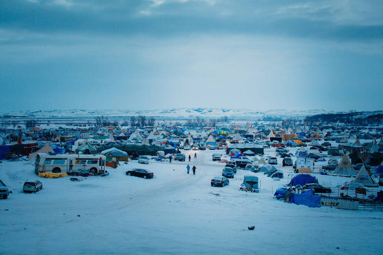 A imagem mostra um grande acampamento em uma área coberta de neve. Há várias tendas e veículos estacionados， com algumas pessoas caminhando entre eles. O céu está nublado， e ao fundo， podem ser vistas colinas cobertas de neve.