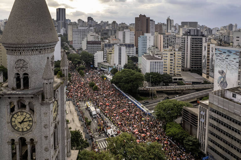 A imagem mostra uma grande manifestação em uma rua de São Paulo， com uma multidão de pessoas reunidas. Ao fundo， é possível ver prédios altos e uma torre de relógio em primeiro plano. A rua está parcialmente bloqueada por veículos， e há árvores ao longo da via. O céu está nublado， sugerindo um clima sombrio.