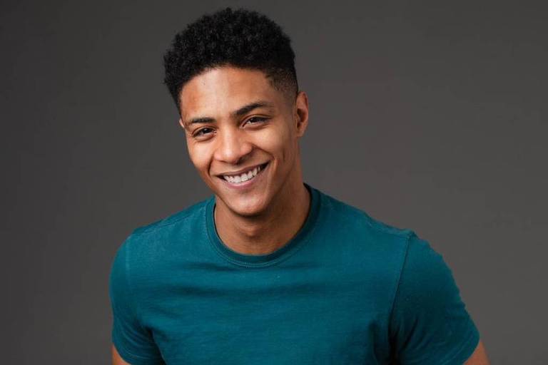 Um jovem sentado em uma cadeira, sorrindo. Ele usa uma camiseta azul-escura e calças pretas. O fundo é cinza, criando um contraste com sua roupa. O jovem tem cabelo curto e encaracolado.