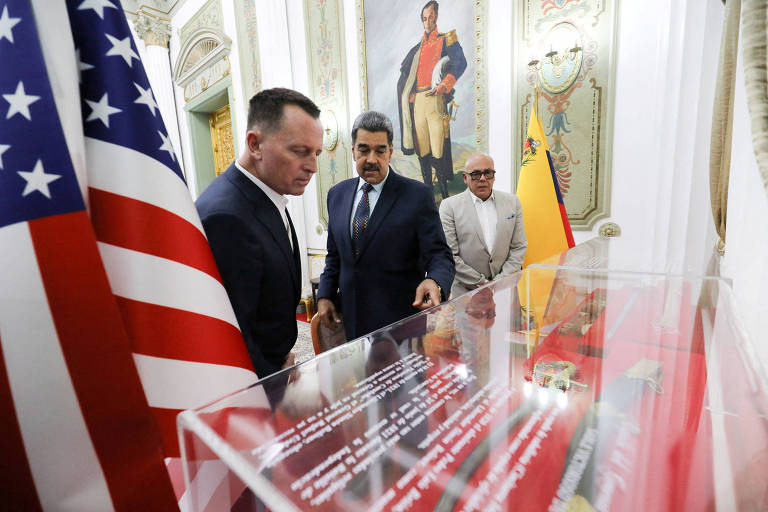 A imagem mostra três homens em um ambiente formal. Um deles， à esquerda， está vestido com um terno escuro e observa atentamente um objeto em uma vitrine. O homem no centro， com um terno azul， também está olhando para a vitrine， enquanto um terceiro homem， à direita， usa óculos e um terno claro. Ao fundo， há uma bandeira dos Estados Unidos e uma pintura de um homem em trajes históricos.