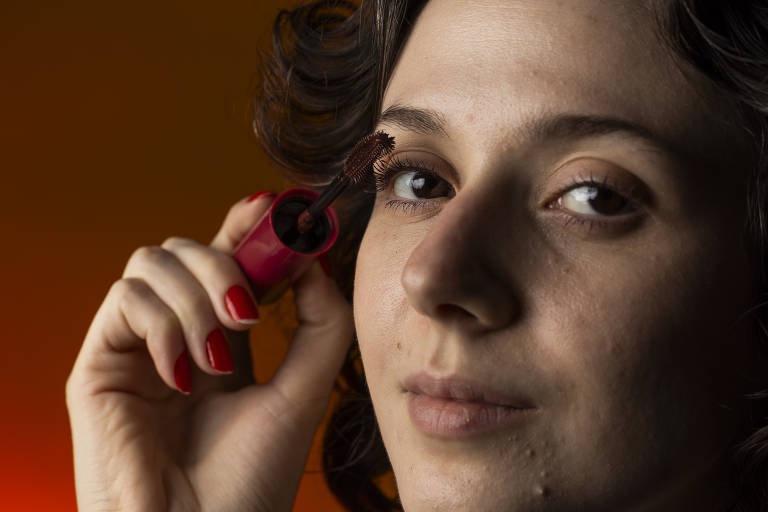 A imagem mostra uma mulher com cabelo cacheado e escuro, segurando um tubo de rímel com uma mão enquanto aplica o produto em seus cílios com a outra. Ela tem uma expressão serena e está olhando para a câmera. O fundo é de uma cor laranja suave.