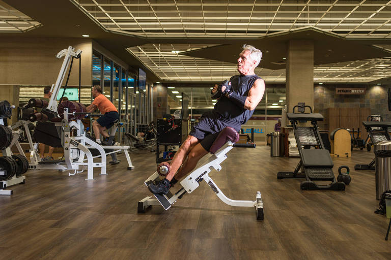 A imagem mostra um homem de cabelos grisalhos， vestindo uma camiseta sem mangas e shorts， realizando um exercício em um equipamento de academia. Ele está inclinado para trás， segurando pesos com as duas mãos. Ao fundo， outras pessoas estão usando diferentes equipamentos de musculação em um ambiente bem iluminado e moderno.