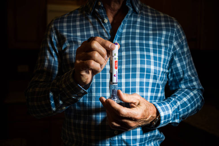 A imagem mostra um homem com uma camisa quadriculada azul e branca， segurando um dispositivo médico em uma mão e com a outra mão apontando para ele. O fundo é escuro， sugerindo que a iluminação é focada no dispositivo. O homem parece estar em uma cozinha.