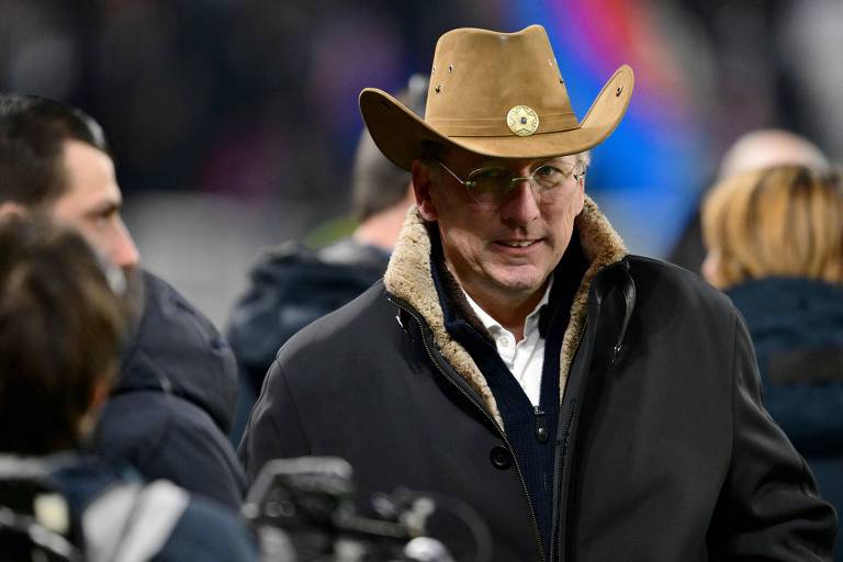 Um homem está em um evento， usando um chapéu de cowboy e um casaco escuro com forro de pele. Ele parece estar sorrindo e interagindo com outras pessoas ao seu redor. O fundo é desfocado， mas há outras pessoas e câmeras visíveis.