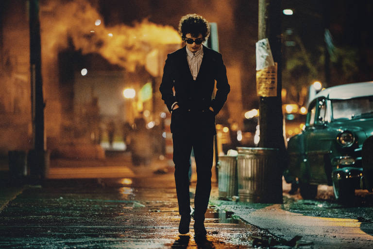 Um homem com cabelo encaracolado e óculos escuros caminha sozinho em uma rua urbana à noite. Ele está vestido com um terno preto e uma camisa branca, com as mãos nos bolsos. Ao fundo, há fumaça e luzes de uma cidade, além de um carro antigo verde estacionado. O ambiente parece molhado, possivelmente devido à chuva.