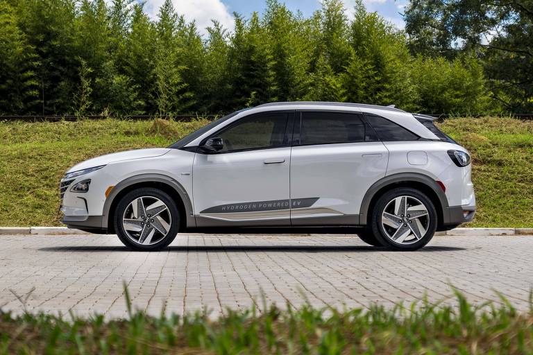 Um carro SUV branco está estacionado em uma estrada de paralelepípedos， com uma vegetação verde ao fundo. O céu está parcialmente nublado， com algumas nuvens brancas. O carro é visto de lado， destacando seu design moderno e rodas de liga leve.