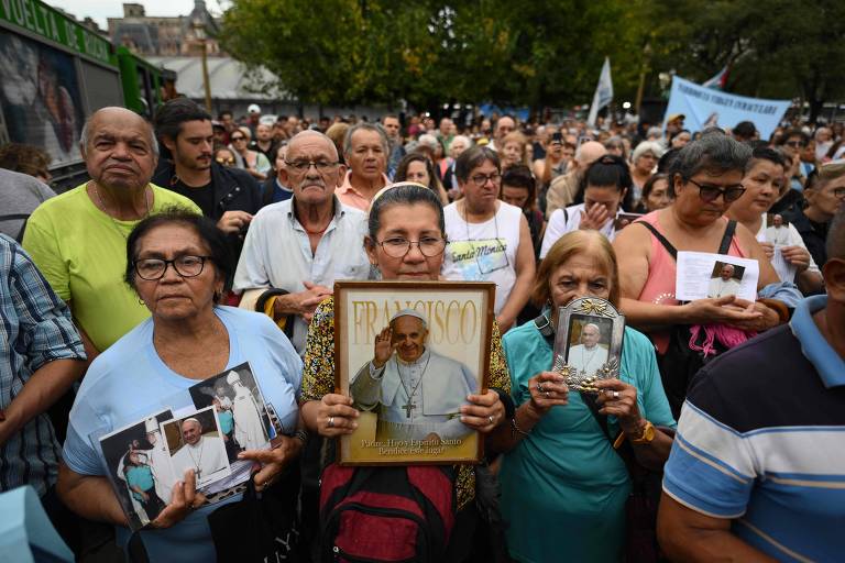 A imagem mostra uma multidão de pessoas em uma manifestação， com foco em um grupo de mulheres na frente segurando fotos do Papa Francisco. Elas estão cercadas por outras pessoas que também seguram imagens e cartazes. O ambiente é ao ar livre， com árvores ao fundo e uma atmosfera de apoio e devoção.