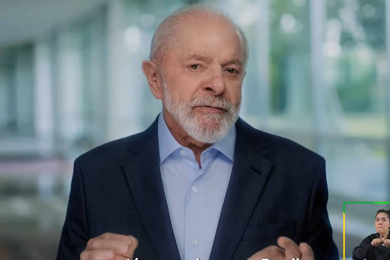 Um homem de cabelos grisalhos e barba， vestido com uma camisa azul clara e um paletó escuro， está falando em um ambiente interno com grandes janelas ao fundo. Ele gesticula com as mãos enquanto se expressa. À direita da imagem， há uma pessoa fazendo linguagem de sinais. Na parte inferior da imagem， está escrito: 039;mas que está mexendo com o Brasil!039;.