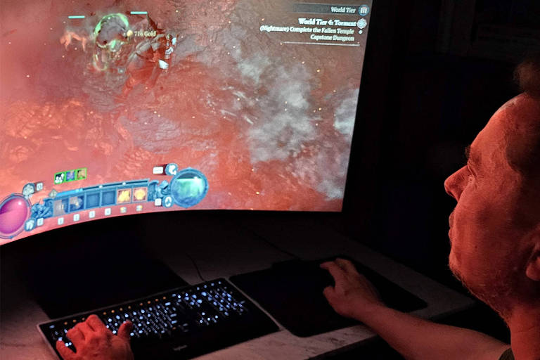 Um homem está jogando em um computador com um monitor curvo grande. Ele está concentrado na tela， que exibe um jogo com gráficos coloridos. O teclado iluminado está à sua frente， e a iluminação ambiente é suave.